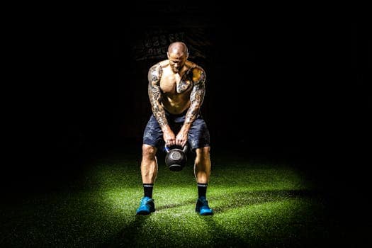 Muscular man with tattoos lifting a kettlebell indoors under a spotlight. Intense fitness and strength training.